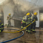 Firefighters in full gear work to extinguish a house garage fire, yellow hoses on a wet driveway as smoke billows from the structure behind a collapsed door frame.