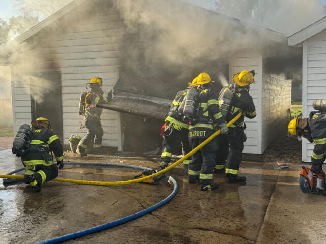 Firefighters in full gear work to extinguish a house garage fire, yellow hoses on a wet driveway as smoke billows from the structure behind a collapsed door frame.