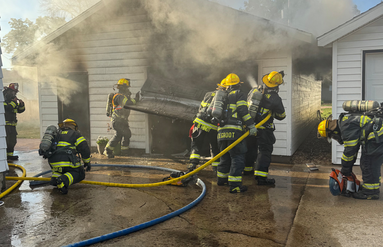 Firefighters in full gear work to extinguish a house garage fire, yellow hoses on a wet driveway as smoke billows from the structure behind a collapsed door frame.