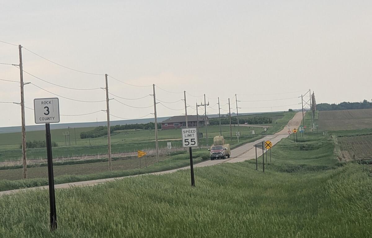 Rural two-lane road through farmland with utility poles, signs, and a distant pickup truck traveling away.
