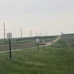 Rural two-lane road through farmland with utility poles, signs, and a distant pickup truck traveling away.