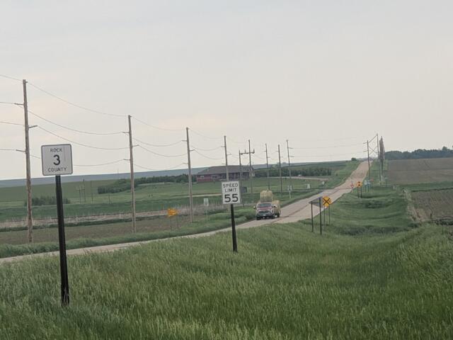 Rural two-lane road through farmland with utility poles, signs, and a distant pickup truck traveling away.