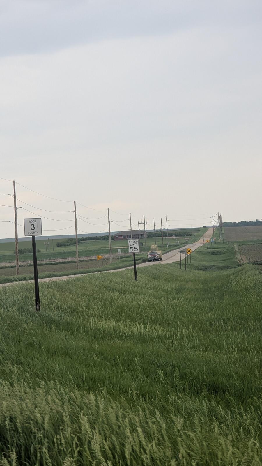 Rural two-lane road through farmland with utility poles, signs, and a distant pickup truck traveling away.