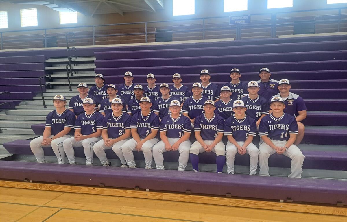 Group photo of a high school baseball team in purple uniforms labeled 'TIGERS' on gym bleachers, with coaches in back row.