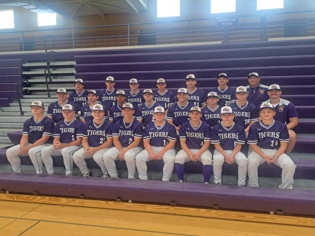 Group photo of a high school baseball team in purple uniforms labeled 'TIGERS' on gym bleachers, with coaches in back row.