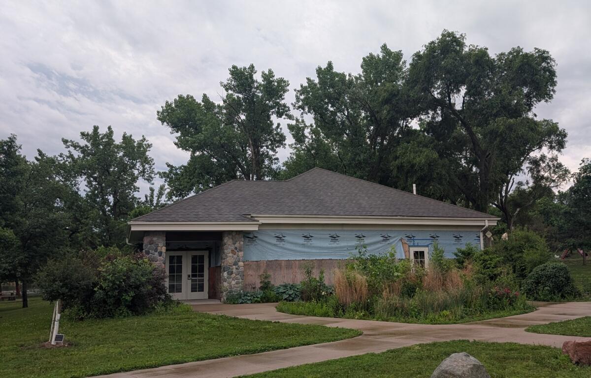 Single-story building under renovation with a blue weather barrier, stone columns at the entrance, and a curved sidewalk in a landscaped area beneath a cloudy sky.