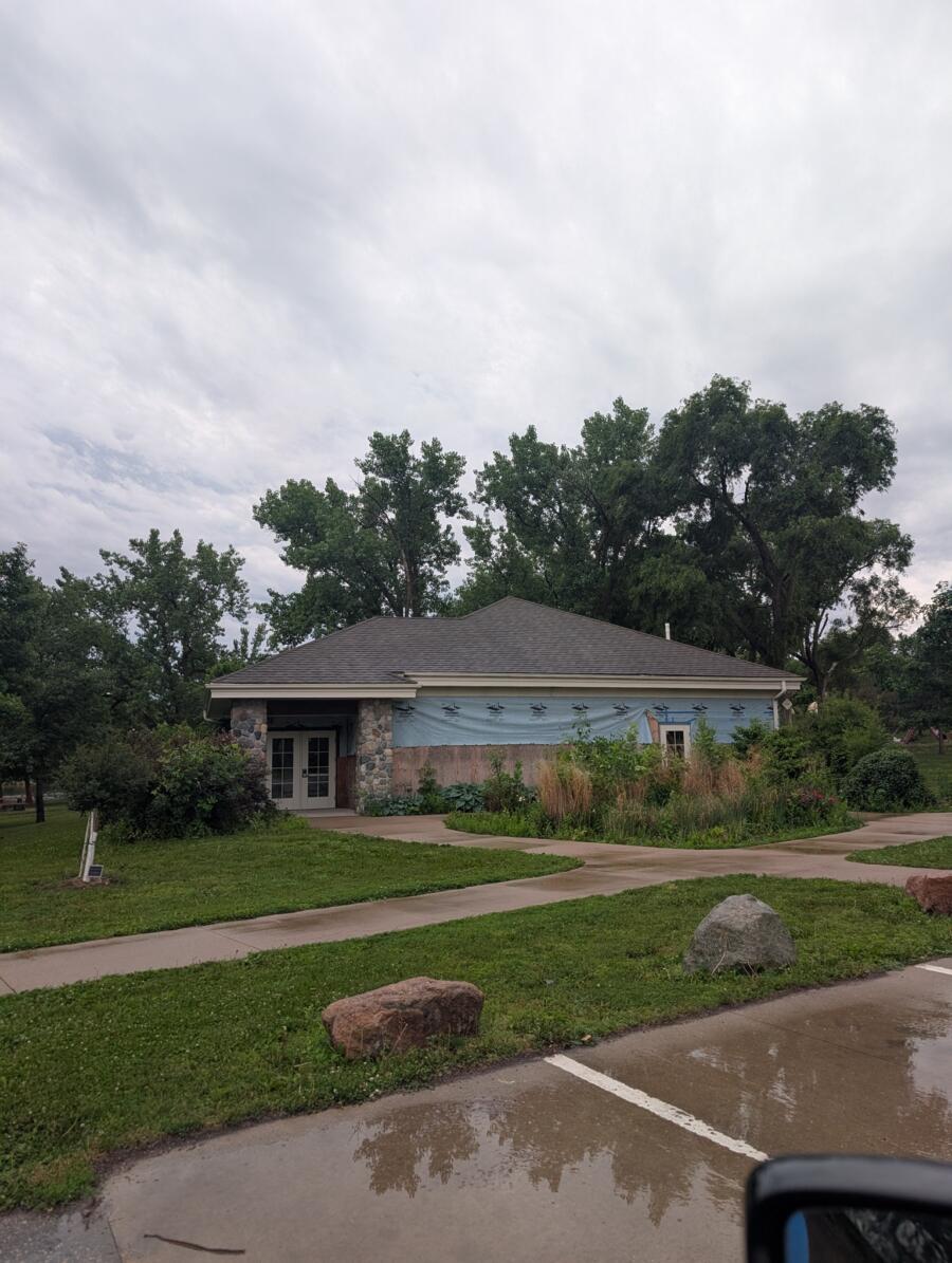 Single-story building under renovation with a blue weather barrier, stone columns at the entrance, and a curved sidewalk in a landscaped area beneath a cloudy sky.