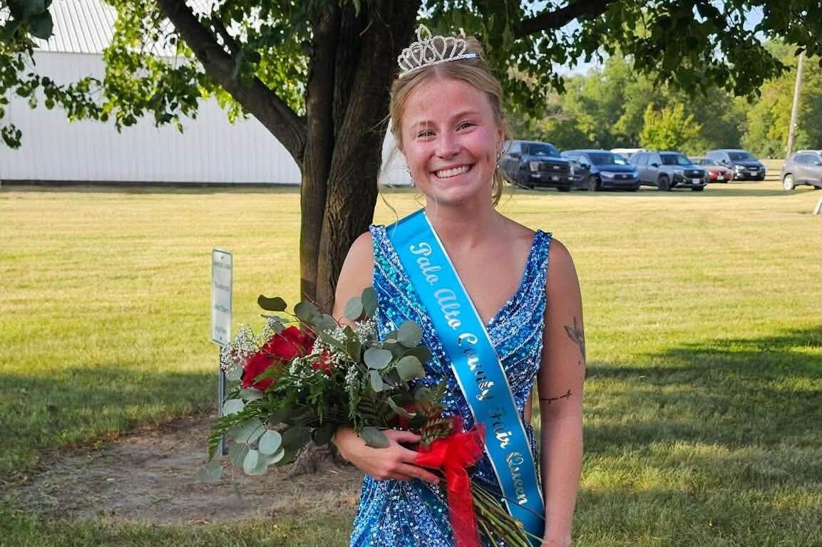Smiling young woman in a blue sequined dress with a tiara, wearing a blue sash and holding a bouquet of red roses outdoors under a tree.
