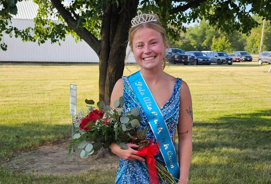 Smiling young woman in a blue sequined dress with a tiara, wearing a blue sash and holding a bouquet of red roses outdoors under a tree.