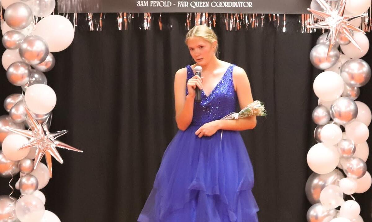 Young woman in a blue sequined gown speaks into a microphone on a decorated stage with silver and white balloons framing the backdrop.