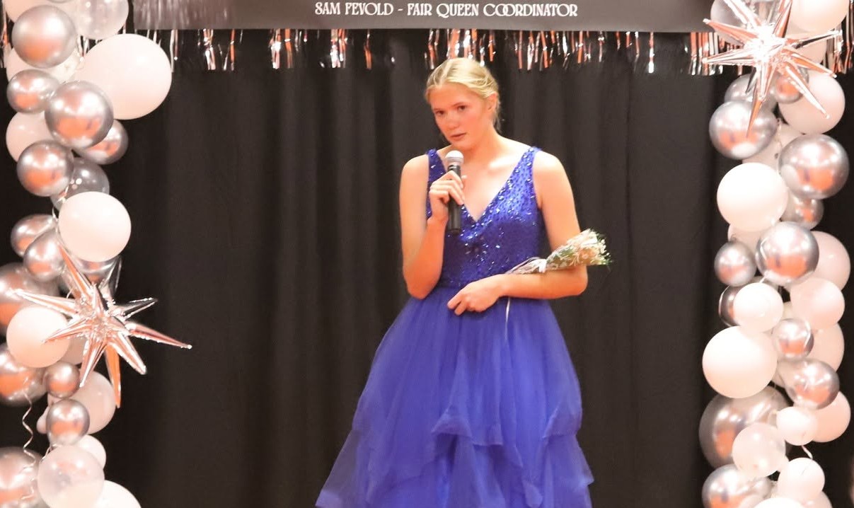 Young woman in a blue sequined gown speaks into a microphone on a decorated stage with silver and white balloons framing the backdrop.