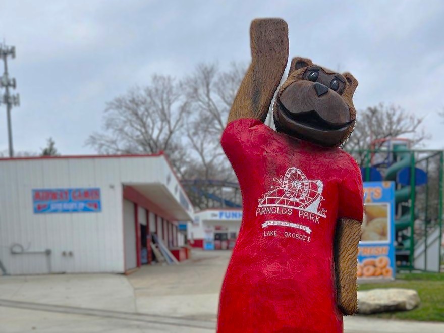 Mascot bear statue in a red shirt at Arnold's Park, raising one arm in the air near a fun center exterior in an amusement park setting.