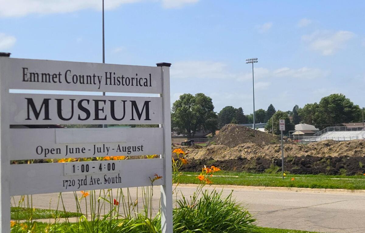 Sign for Emmet County Historical Museum with open hours text and flowers in foreground; construction site and blue sky in background.