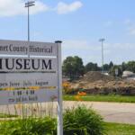 Sign for Emmet County Historical Museum with open hours text and flowers in foreground; construction site and blue sky in background.