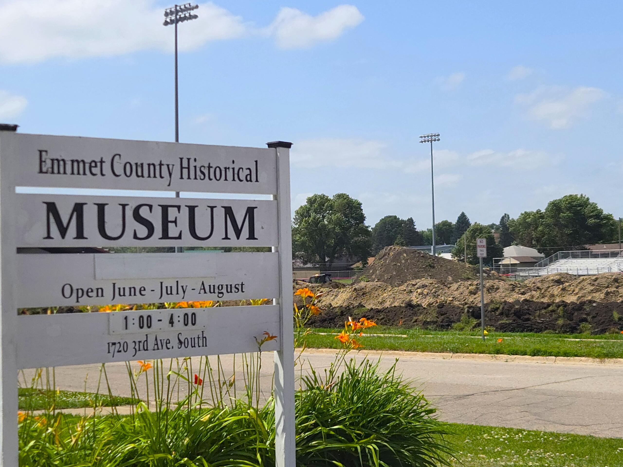 Sign for Emmet County Historical Museum with open hours text and flowers in foreground; construction site and blue sky in background.