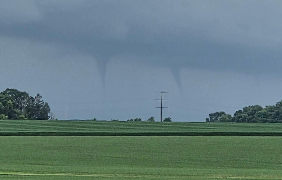 Tornado funnel extending from a dark storm cloud over a flat green field with a lone power pole in the distance.