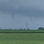 Tornado funnel extending from a dark storm cloud over a flat green field with a lone power pole in the distance.