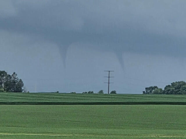 Tornado funnel extending from a dark storm cloud over a flat green field with a lone power pole in the distance.
