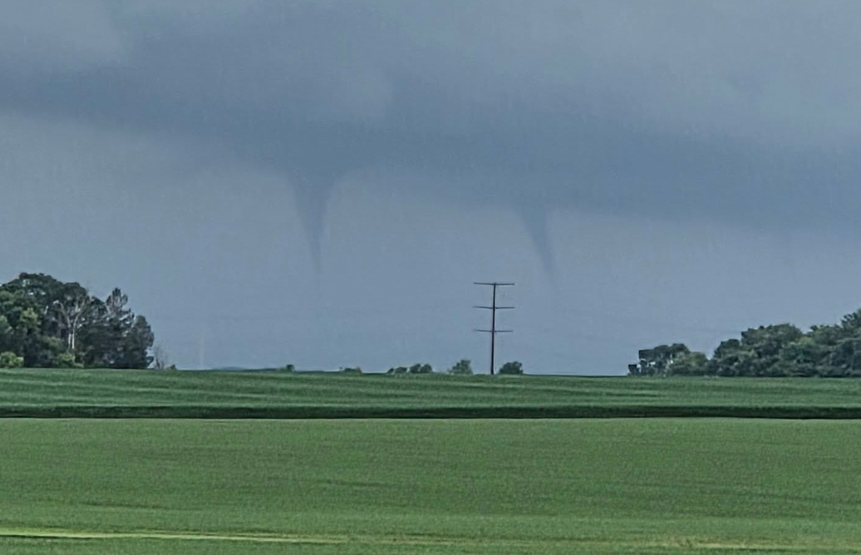 Tornado funnel extending from a dark storm cloud over a flat green field with a lone power pole in the distance.