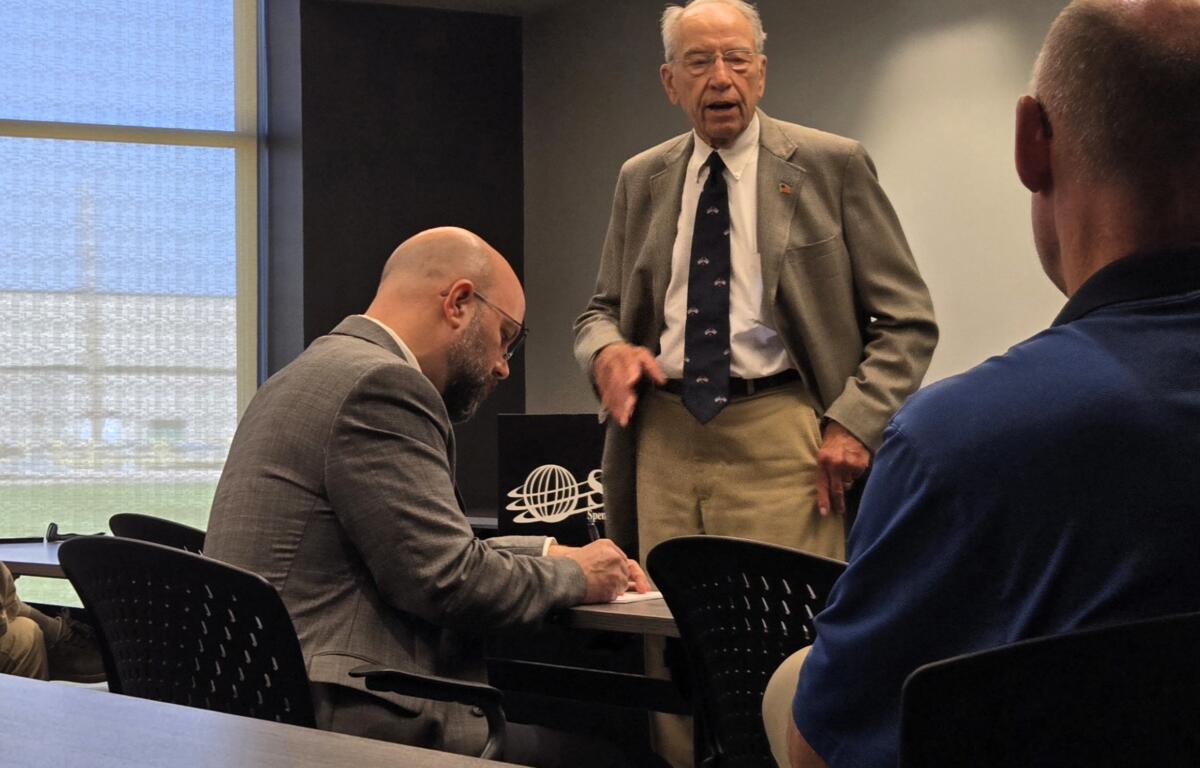 Elderly man in a gray suit speaks to colleagues during a business meeting in a conference room.
