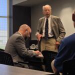 Elderly man in a gray suit speaks to colleagues during a business meeting in a conference room.