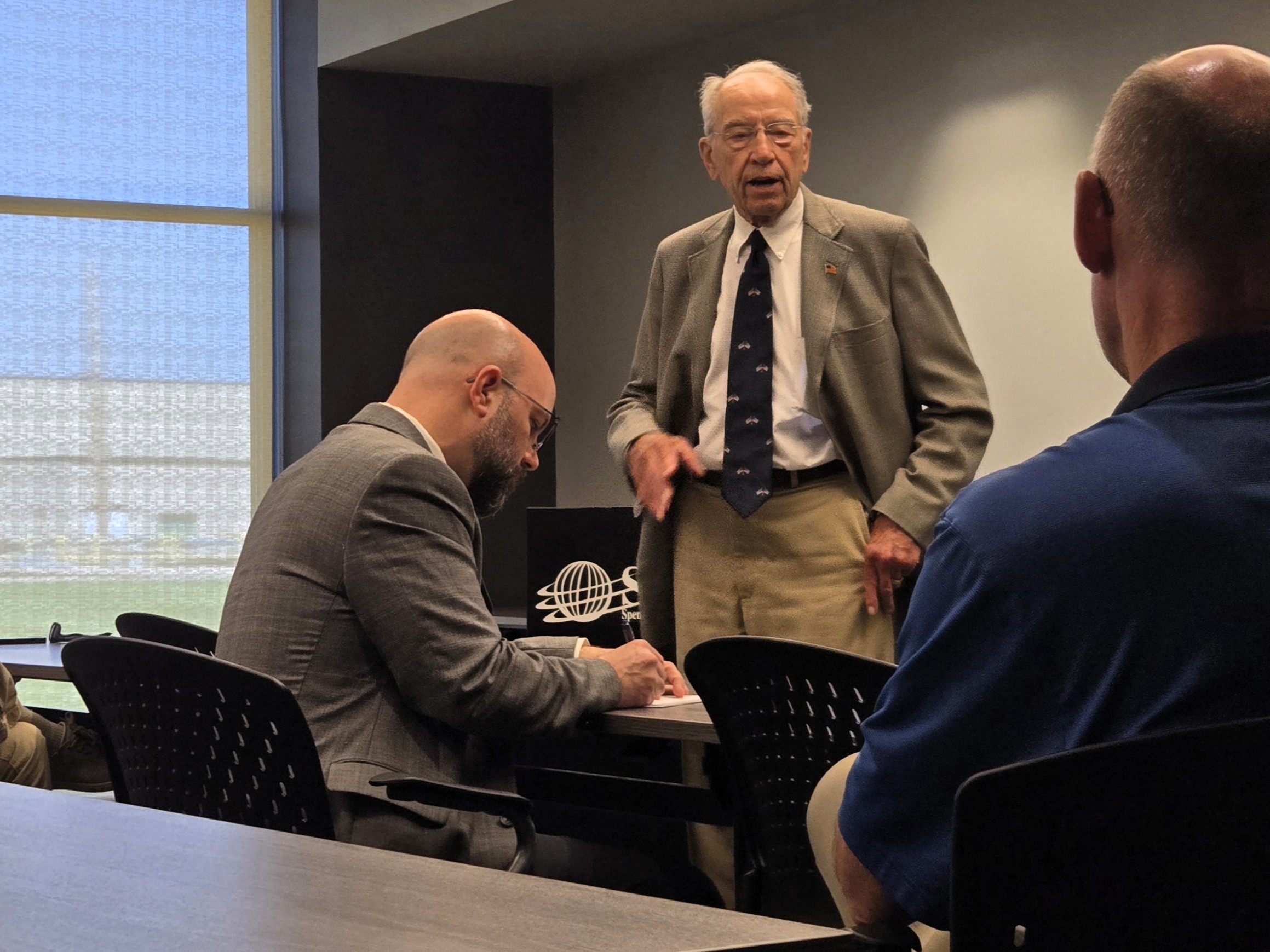 Elderly man in a gray suit speaks to colleagues during a business meeting in a conference room.
