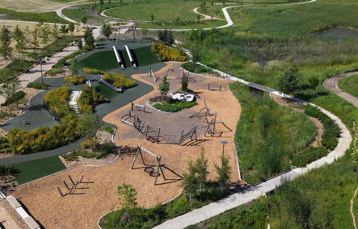 Aerial view of a modern playground with two tall tubular slides, climbing frames, and a wood-chip ground cover, surrounded by winding paths and greenery in a park.