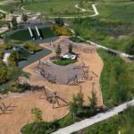 Aerial view of a modern playground with two tall tubular slides, climbing frames, and a wood-chip ground cover, surrounded by winding paths and greenery in a park.