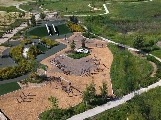 Aerial view of a modern playground with two tall tubular slides, climbing frames, and a wood-chip ground cover, surrounded by winding paths and greenery in a park.