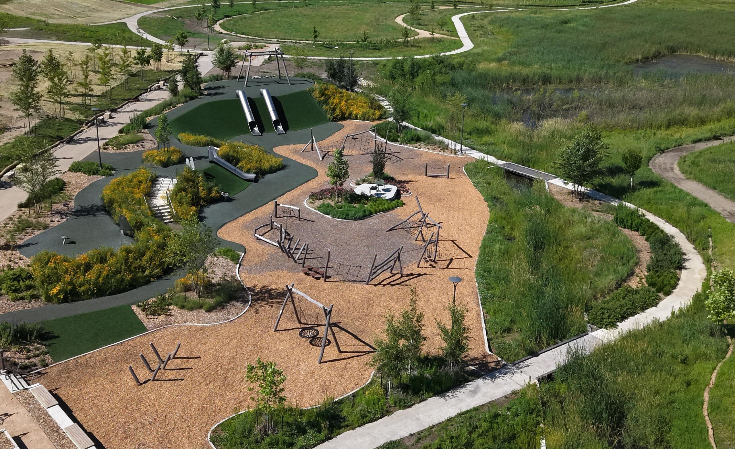 Aerial view of a modern playground with two tall tubular slides, climbing frames, and a wood-chip ground cover, surrounded by winding paths and greenery in a park.