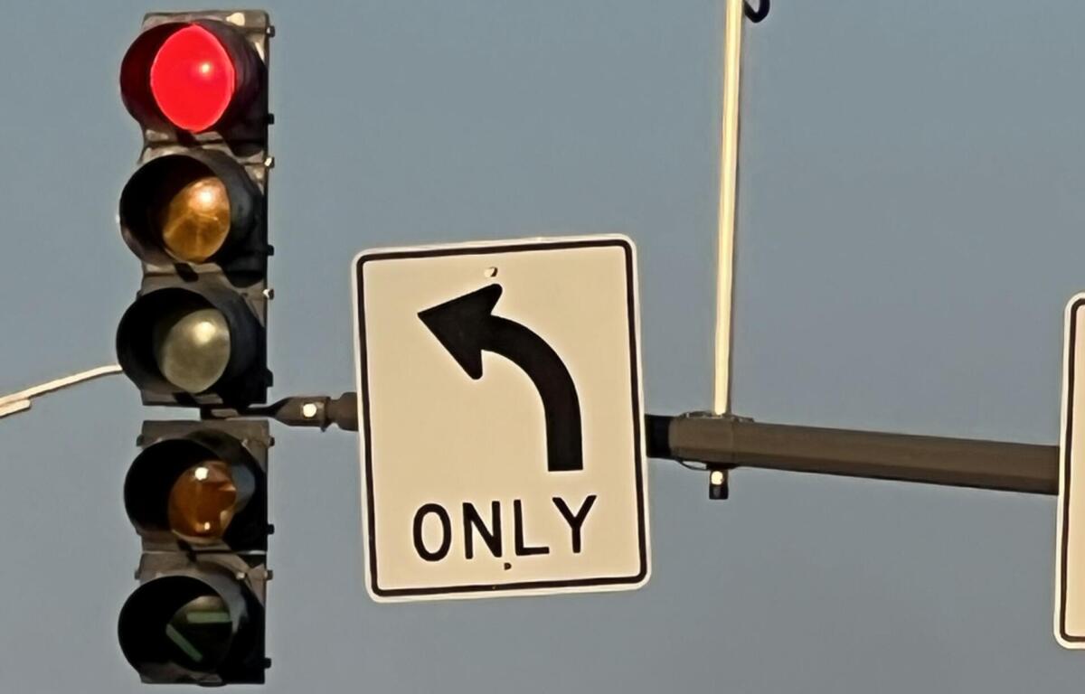 Red traffic light illuminated while a left-turn-only sign and a surveillance camera are visible on a pole against a blue sky.