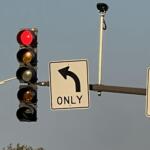 Red traffic light illuminated while a left-turn-only sign and a surveillance camera are visible on a pole against a blue sky.