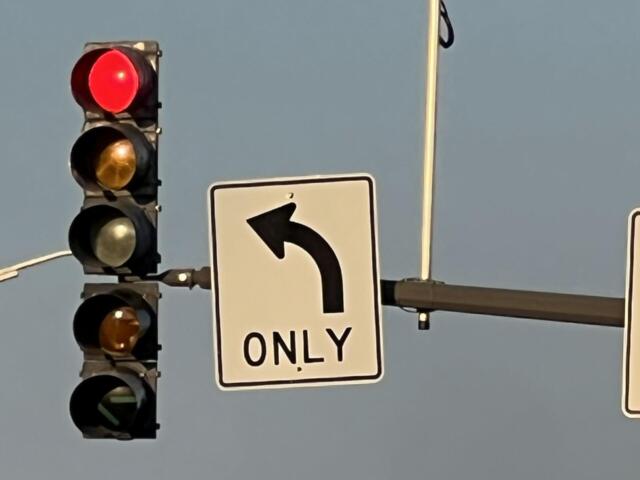 Red traffic light illuminated while a left-turn-only sign and a surveillance camera are visible on a pole against a blue sky.