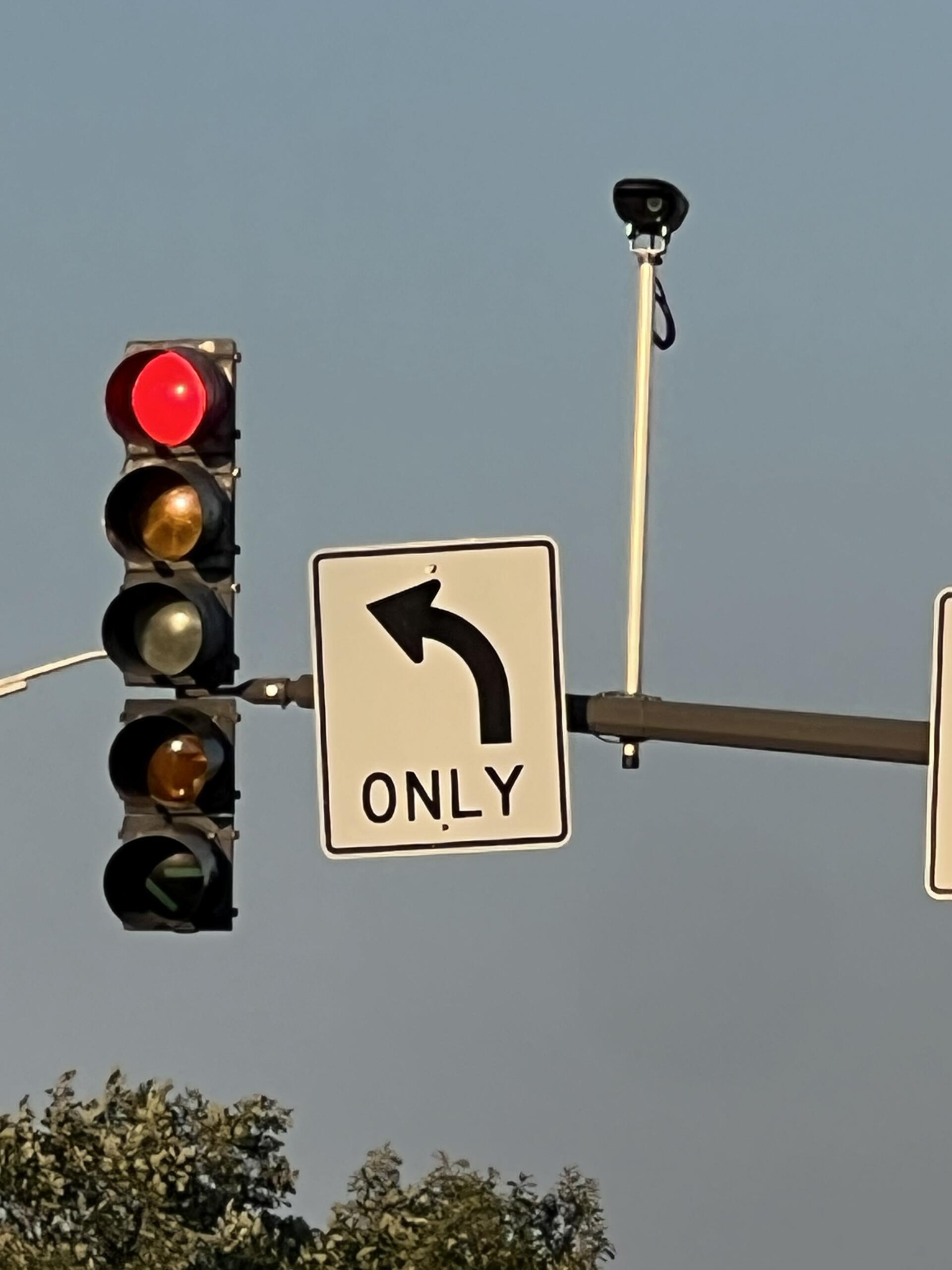 Red traffic light illuminated while a left-turn-only sign and a surveillance camera are visible on a pole against a blue sky.