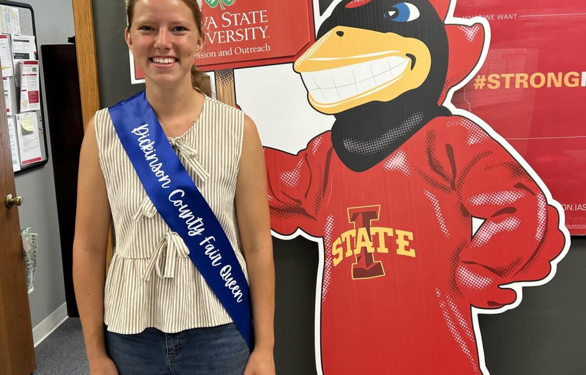 Smiling girl wearing a crown and blue sash reading 'Johnson County Fair Queen' poses beside a large red cartoon mascot.