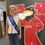 Smiling girl wearing a crown and blue sash reading 'Johnson County Fair Queen' poses beside a large red cartoon mascot.