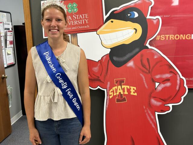 Smiling girl wearing a crown and blue sash reading 'Johnson County Fair Queen' poses beside a large red cartoon mascot.
