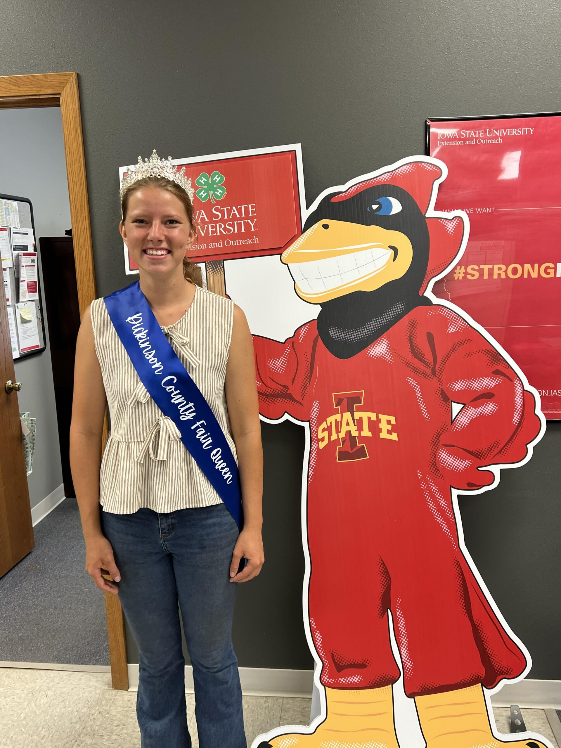 Smiling girl wearing a crown and blue sash reading 'Johnson County Fair Queen' poses beside a large red cartoon mascot.