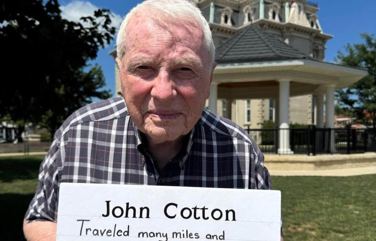 Older man in a plaid shirt holds a sign about traveling all towns in Iowa, outdoors with a historic building in the background.