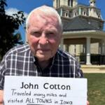 Older man in a plaid shirt holds a sign about traveling all towns in Iowa, outdoors with a historic building in the background.
