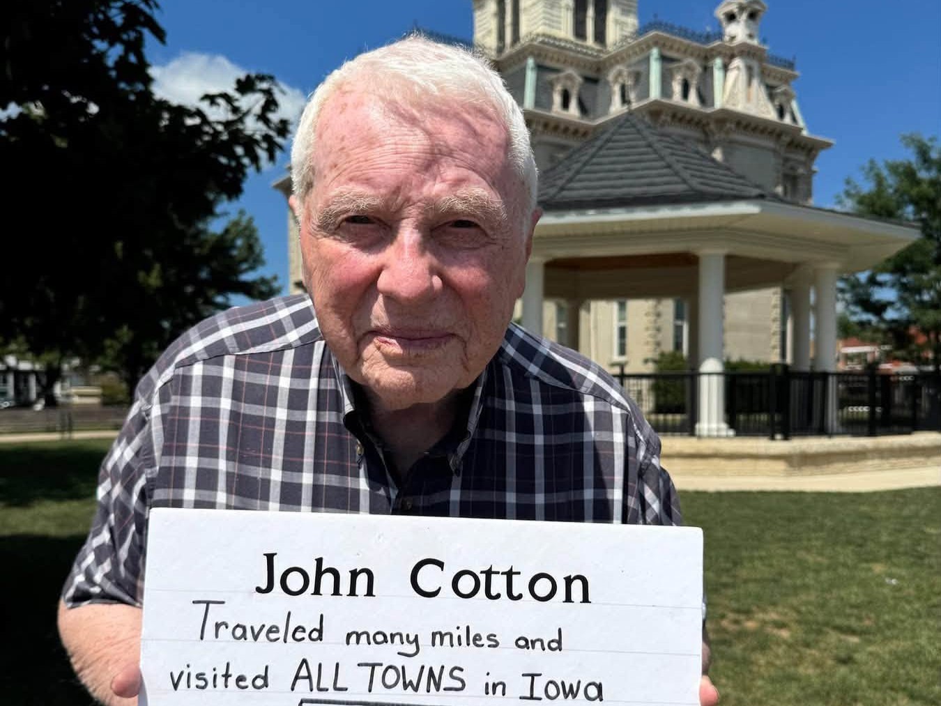 Older man in a plaid shirt holds a sign about traveling all towns in Iowa, outdoors with a historic building in the background.