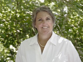 Portrait of a smiling woman in a white blouse with a pearl necklace, standing in a sunlit garden with green leaves in the background