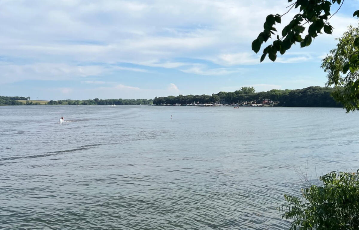 Wide lake scene with a small motorized boat speeding across water and a tree-lined shoreline in the distance, under a blue sky.