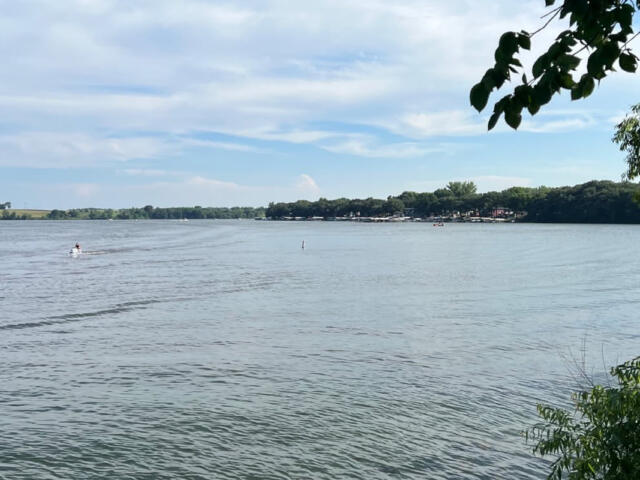 Wide lake scene with a small motorized boat speeding across water and a tree-lined shoreline in the distance, under a blue sky.