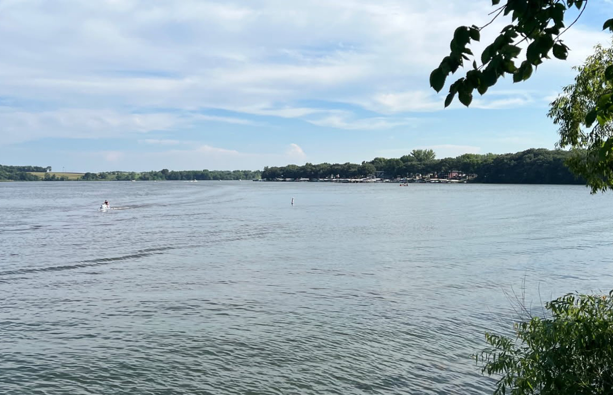 Wide lake scene with a small motorized boat speeding across water and a tree-lined shoreline in the distance, under a blue sky.