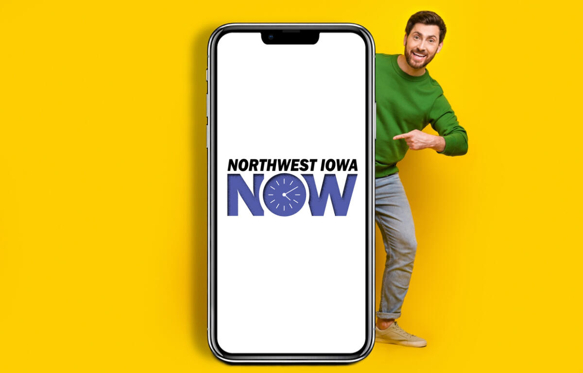 Smiling man in a green sweater peeks from the right behind a large smartphone displaying the Northwest Iowa NOW logo on a white screen against a yellow background.