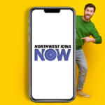 Smiling man in a green sweater peeks from the right behind a large smartphone displaying the Northwest Iowa NOW logo on a white screen against a yellow background.