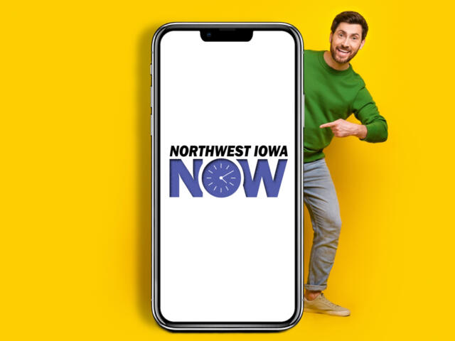 Smiling man in a green sweater peeks from the right behind a large smartphone displaying the Northwest Iowa NOW logo on a white screen against a yellow background.
