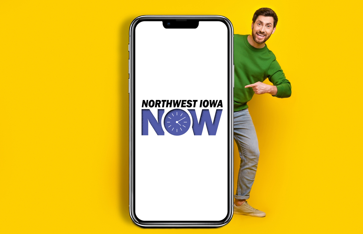 Smiling man in a green sweater peeks from the right behind a large smartphone displaying the Northwest Iowa NOW logo on a white screen against a yellow background.