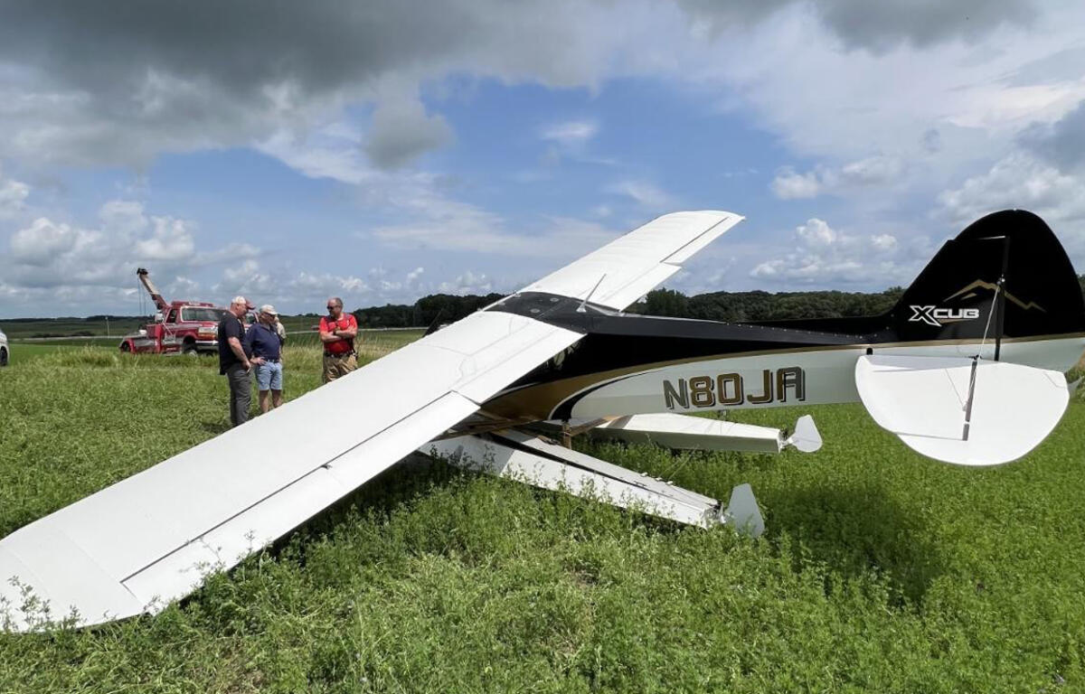 Small white light aircraft down in a grassy field with several men examining the scene; a red tow truck is in the background.
